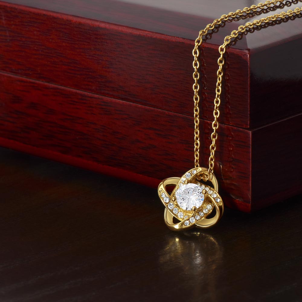 Gold love knot pendant necklace with sparkling crystal center — delicate chain draped over mahogany gift box, graduation or daughter jewelry gift